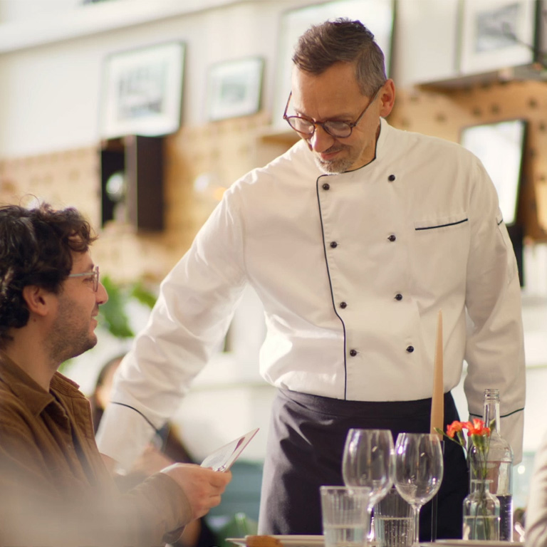 Ein Gastronom steht neben einem Tisch und unterhält sich mit dem sitzenden Gast